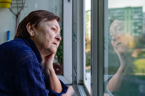 Portrait of a sad elderly woman looking out the window at home. A lonely elderly person looking out of a window