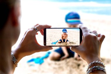 Küçük oğlunun cep telefonuyla fotoğrafını çeken bir kadın. Çocuk kumsaldaki kumda oynuyor. Akıllı telefonun kamerası..