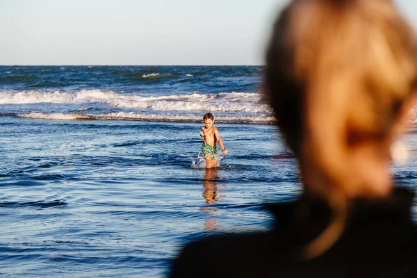 Güneşli bir yaz gününün günbatımında anne onu dikkatle izlerken bir çocuk deniz dalgalarında eğleniyor..