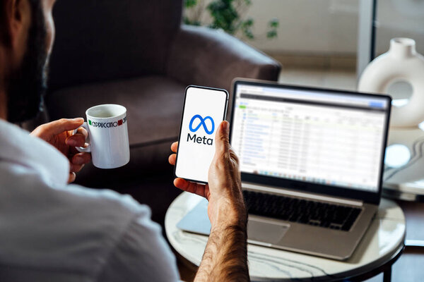 Man holding a smartphone showing Meta logo on mobile phone screen. Laptop with Facebook business manager on background. Rosario, Argentina - June 20, 2024.