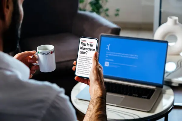 Mavi ekranı ya da dizüstü bilgisayarının BSOD 'unu tamir etmek için cep telefonuyla bilgi arayan bir adam. Hatalı işletim sistemi hatası.