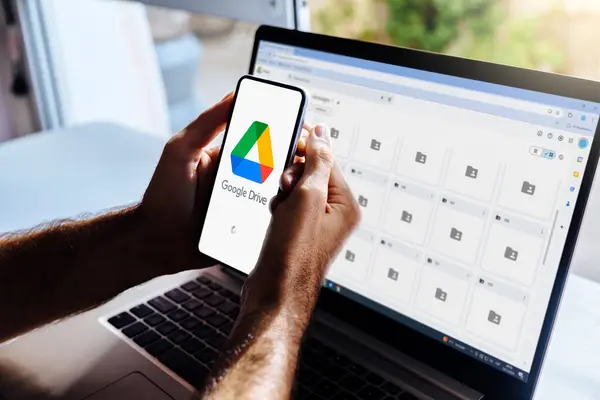 Cep telefonu ve bilgisayarda Google Drive. Ekranda bulut depolama uygulaması olan bir akıllı telefon tutan adam. Arka plandaki dizüstü bilgisayar Google sürücüsü dizinlerini gösteriyor. Rosario, Arjantin - 26 Temmuz 2024.