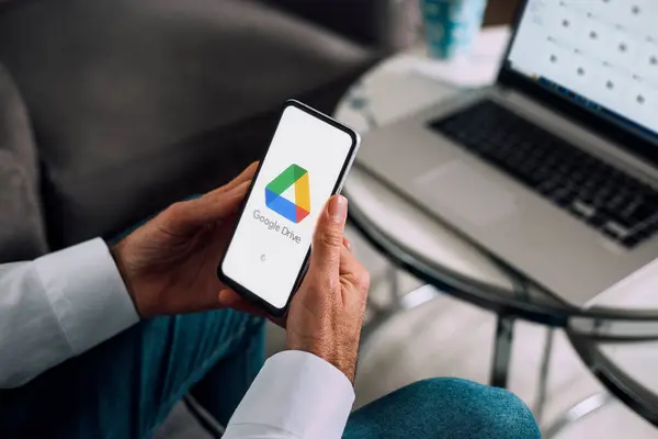Cep telefonu ve bilgisayarda Google Drive. Ekranda bulut depolama uygulaması olan bir akıllı telefon tutan adam. Arka plandaki dizüstü bilgisayar Google sürücüsü dizinlerini gösteriyor. Rosario, Arjantin - 26 Temmuz 2024.