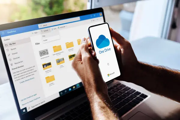 Cep telefonu ve bilgisayarda bir sürücü. Ekranda Microsoft bulut depolama uygulaması olan bir cep telefonu tutan adam. Arka plandaki dizüstü bilgisayar One Drive klasörlerini gösteriyor. Rosario, Arjantin - 27 Ocak 2025