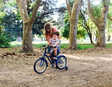 Annem küçük oğluna bisiklet sürmeyi öğretiyor. Orman parkında mavi bir bisiklete binen küçük bir çocuk ve annesi ona yardım etti. Destek tekerlekleri olan bisiklet.