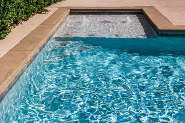 A rectangular new swimming pool with tan concrete edges in the fenced backyard of a new construction house.