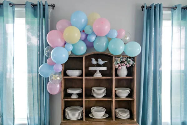 A pastel balloon arch for baby shower decorations with awindows and aqua curtains.