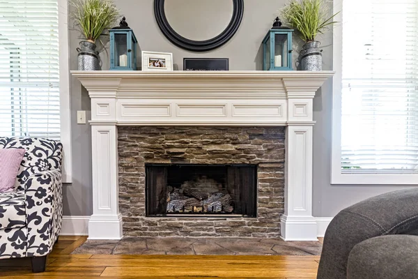 A white wood fireplace in a new home with windows and a chair in a living room.