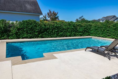 A rectangular new swimming pool with tan concrete edges in the fenced backyard of a new construction house.