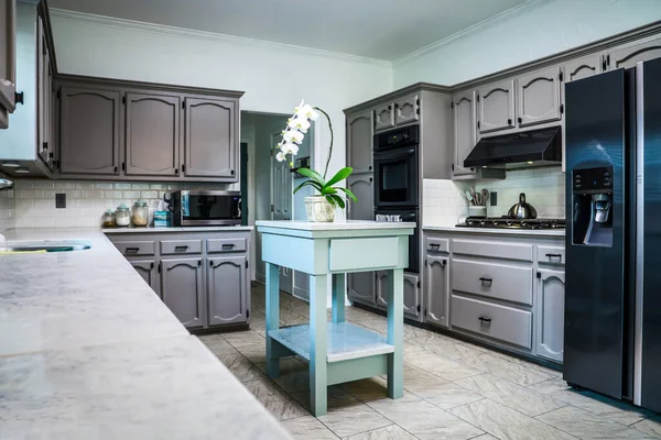 A renovated kitchen in an older home with painted gray cabinets, marble countertops, a small portable island and a tiled floor.