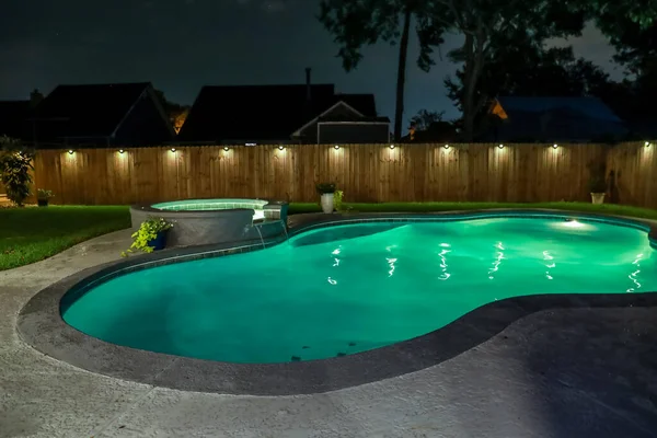 A backyard swimming pool and jacuzzi hot tob at night with solar lights around the fence for privacy and illumination.