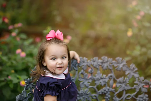 A toddler little girl with a navy dress and pink bow is sitting on an iron vintage bench outside.