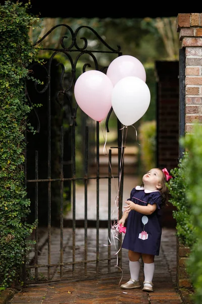 Little toddler girl with pink birthday balloons outside near an iron fence with a navy dress and pink hair bow. She is looking up at the balloons.