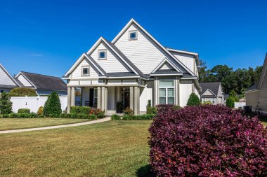 The front view of a new construction cottage craftsman style white house with a triple pitched roof with a sidewalk, landscaping and curb appeal.