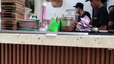 Tezgahın arkasında çalışan samimi baristaların fotoğrafıyla hareketli bir kafenin enerjisini yakalayın. Tabaklar, renkli bardaklar ve çeşitli malzemeler personel olarak çalışma alanını dolduruyor.