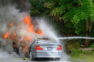 Saint John, NB, Kanada - 18 Ağustos 2022: Bir araba alevler içinde kaldı. Köpüklü bir su akıntısı sağdan ateşle savaşıyor. Arka planda ağaçlar var. Çok duman var..