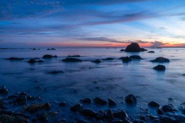 Gün batımında, volkanik kayalarla şafak vakti sahilde görülmüş. Bulutlar, berrak mavi gökyüzü, fantezi manzarası.