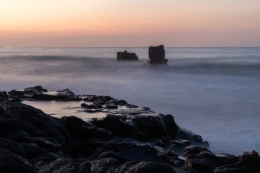 Fotoğraf, milyonlarca yıl önce, alacakaranlıkta, denizin üzerinde iki lav kayasının yükseldiği antik bir lav kayasındaki deniz manzarasını gözler önüne seriyor. Sıcak fotoğraf rengi. Yumuşak odak.