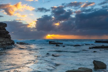 Autumn sunrise with the sun's rays beaming through the clouds over the sea and rocks at The Skillion in Terrigal, NSW, Australia. clipart