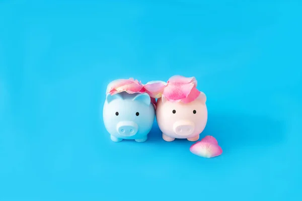 A blue and a pink piggy bank with rose petals on heads against blue background. Minimal surreal concept for Valentine or wedding card or banner. Design for editorial on love and money
