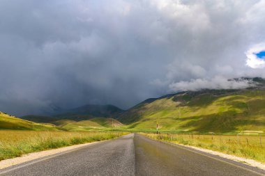 La strada che taglia l'altipiano di Castelluccio di Norcia in una giornata nuvolosa d'estate durante la fioritura delle lenticchie, Umbria, Italia