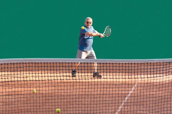 Active senior enjoys sports while playing tennis. Elderly tennis player playing ball on red clay court. Vitality, sports activity, healthy lifestyle in old age, aging youthfully concept