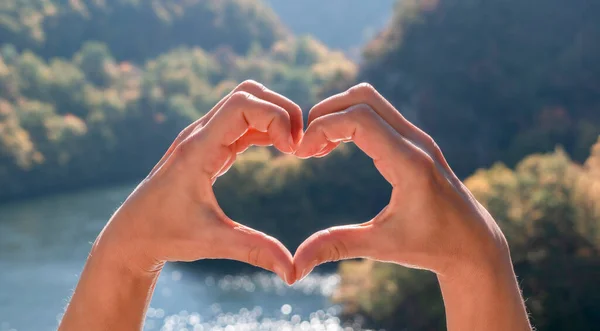 Love nature, travel, Earth day concept. Shape of heart, made by women hands on backgroubd of beautiful natural landscape - water surface of mountain lake and forested green mountains. Panoramic banner