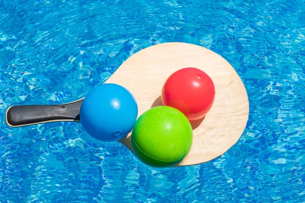 Summer play and vacation fun, water games, beach activity and sport concept. Multi-colored balls on beach tennis racket float in blue water of swimming pool.