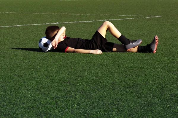 Cute boy soccer player lies on the grass of a football field, resting after training. Child soccer player and a soccer ball. Sport concept. Kids football school, tournament. Background. Copy space