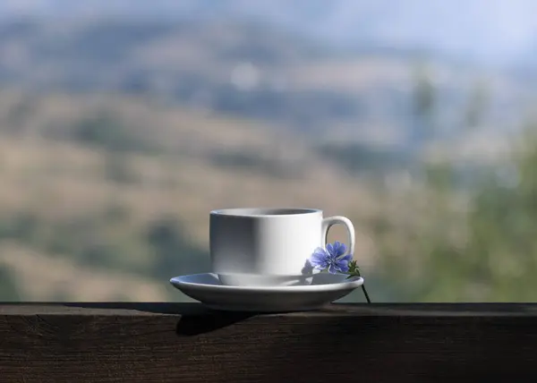 Summer serene morning among nature. Cup of coffee, blue delicate flower on wooden railing of rural house and blurred background of mountain valley. Break, breakfast, slow simple life outdoor concept