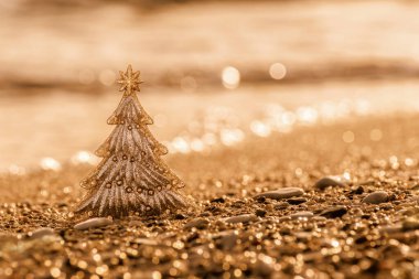 Altın kumsaldaki Noel ağacı ve deniz dalgaları üzerindeki bulanık parıltı. Parlak, altın sarısı bir yeni yıl ya da Noel arkaplanı, tebrik kartı, fotokopi alanı. Sıcak ülkelerde kış tatillerini kutlamak