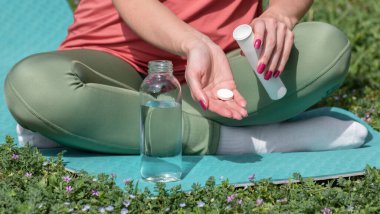 Elinde çözünür tablet tutan genç bir kadın, coşkulu spor içecekleri hazırlıyor, açık hava spor egzersizleri sırasında yoga minderinde oturuyor. Besin takviyeleri veya vitamin eksikliğinin önlenmesi