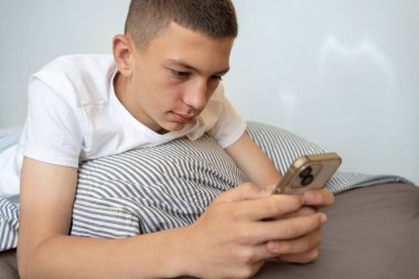 Genç çocuk akıllı telefon kullanırken yatakta dinleniyor. Rahat genç çocuk yatakta uzanıyor, telefona bakıyor, videolar izliyor, mesajlar okuyor, ya da sosyal medyada geziniyor.