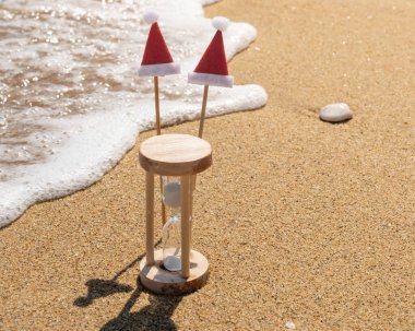 Yaratıcı Noel veya Yeni Yıl kompozisyonu, kumsalda kum saati, çubuklarda süslü Noel Baba şapkaları ve yuvarlanan deniz dalgaları. Mutlu Noeller ve yeni yıl tatili kutlaması zamanı.