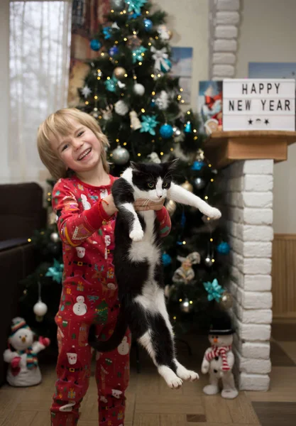 クリスマスの赤いパジャマの明るい少年はクリスマスツリーの前の家に彼の腕の国内の黒と白の猫を保持する ペットと一緒に幸せな居心地の良い家族の冬の休日の概念 — ストック写真