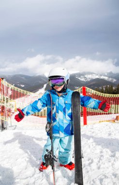 Anaokulu çocuğu, kasklı, mavi tulumlu, dağların tepesinde kar üzerinde kayak botları olan, kayak yapan. Çocuklar için kışın aktif eğlence, spor eğitimi. Sağlıklı bir yaşam tarzı. Neşe. anın tadını çıkar