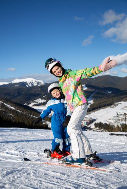 Küçük oğlan ve genç kız, erkek ve kız kardeş, kayak merkezinde dinleniyorlar, dağ yamacında kayakların üstünde neşeyle yan yana duruyorlar. Aile için kış sporları. Mevsimlik kış sevinçleri