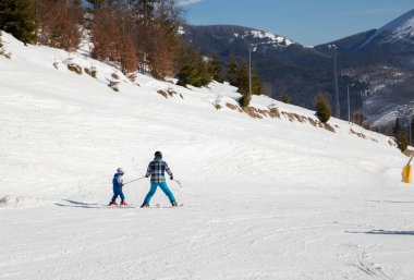 Bir çocuğun ve sıcak tulumlu bir yetişkinin arkasında, kayak yaparken. Dağlardaki karlı bir yamaçta kayak dersi, bir eğitmen olan babamla spor yapmak. Kayak eğitimi. Spor eğitimi. Aktif kış tatili
