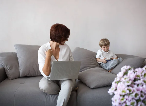 mom and son with digital gadgets are sitting on the sofa in the living room. unrecognizable woman works at home on a laptop, next to a child plays on the phone. concept of modern family life