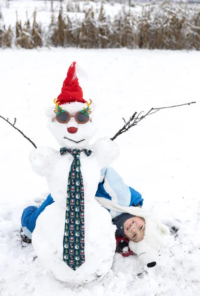 Ребенок Лежит Снегу Весело Смотрит Снеговика Шляпе Санты Карнавальные Очки — стоковое фото