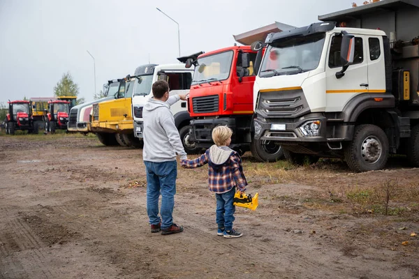 man and a boy, father and son, stand with their backs in front of large trucks. The child\'s passion for cars, dad and child look at the cars in the parking lot with interest