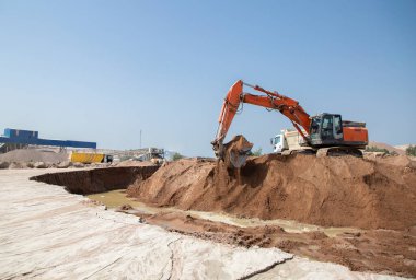 Turuncu sürüngen kazıcı, güneşli bir günde yeraltı suyundan taşan toprak tepesinde çalışıyor. İnşaat alanındaki toprak işleri, iş akışı. İnşaat malzemelerinin kirası ve satışı