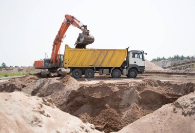 Turuncu sürüngen kazıcı dünyayı sarı damperli kamyonun içine boşaltıyor. İnşaat alanında toprak işleme sürecinde çalışıyorum. İnşaat aracına toprak yükleme ve taşıma.