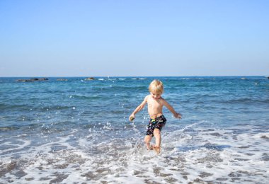 4-5 yaşlarında mutlu bir çocuk, sahildeki deniz suyunda koşuyor. Çocuk sıcak bir yaz gününde mutlu bir şekilde eğleniyor. Sağlıklı yaşam tarzı ve eğlence kavramı. Mutlu çocukluk, merhaba yaz