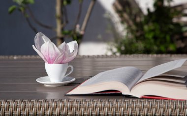 cup with a saucer on the table, there is a large magnolia flower in it, an open book. Good spring morning. Rest time and drink coffee. Read a book with pleasure