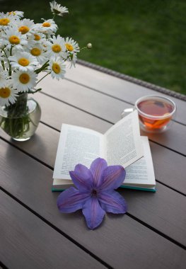 bouquet of daisies, a transparent cup of fragrant tea, an open book on a table in a summer garden. reading books, breakfast, outdoor recreation. Rest and tea drinking. Read with pleasure