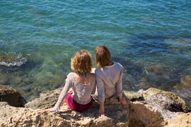 two adult girls sit with their backs on the rocks against the backdrop of the turquoise sea. Mom and adult daughter, girlfriends, sisters on vacation, enjoy the moment