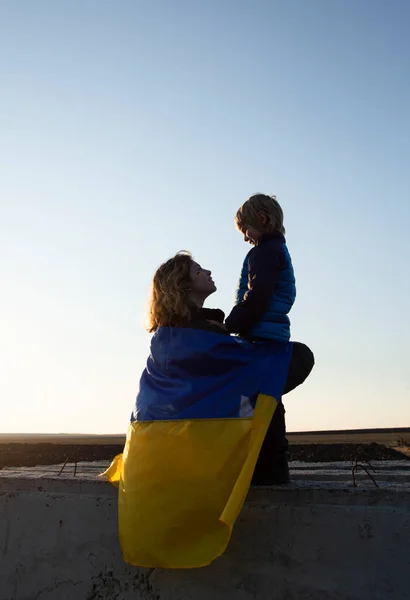 little boy and a young woman are sitting with their backs at sunset against the sky with a blue and yellow Ukrainian flag behind them. Family, refugees, unity, support, love. stand with Ukraine