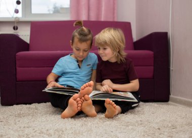 2 barefoot preschool boys are looking at and reading a book together. International Children's Book Day. entertainment for children, interesting education. Two brothers, two friends. Focus on feet