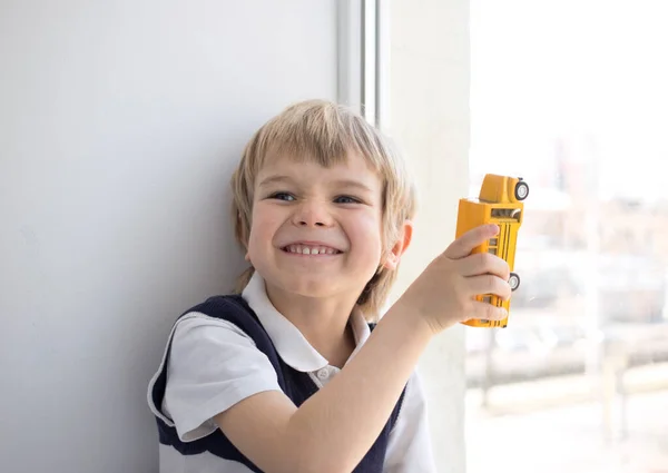 Positive cheerful boy, future schoolboy with a toy yellow school bus near the window. Daylight. The concept of learning, education, soon to school, junior schoolchild